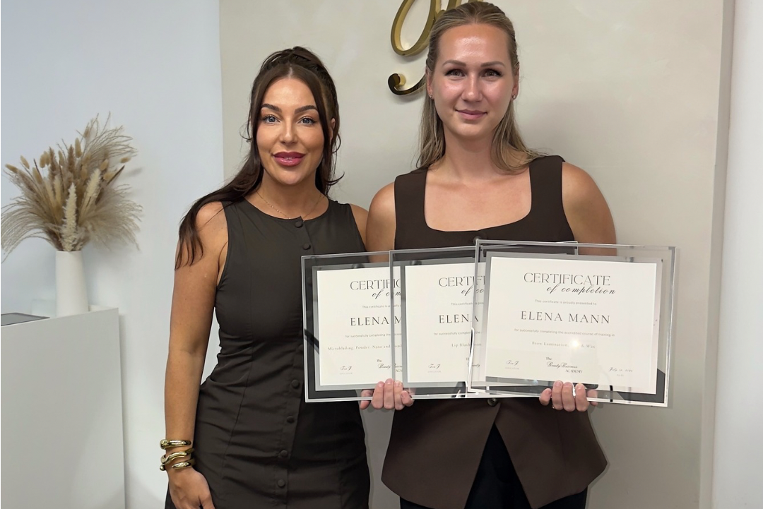 Two women standing indoors and holding three framed certificates. The woman on the left wears a sleeveless dark brown mini dress, and the woman on the right wears a dark brown sleeveless top and blazer over dark pants.